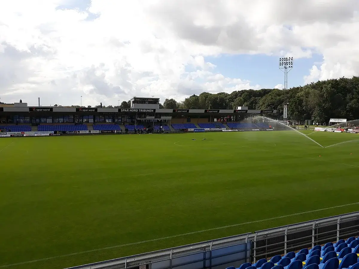 A football stadium with blue seats and sprinklers watering the pitch.