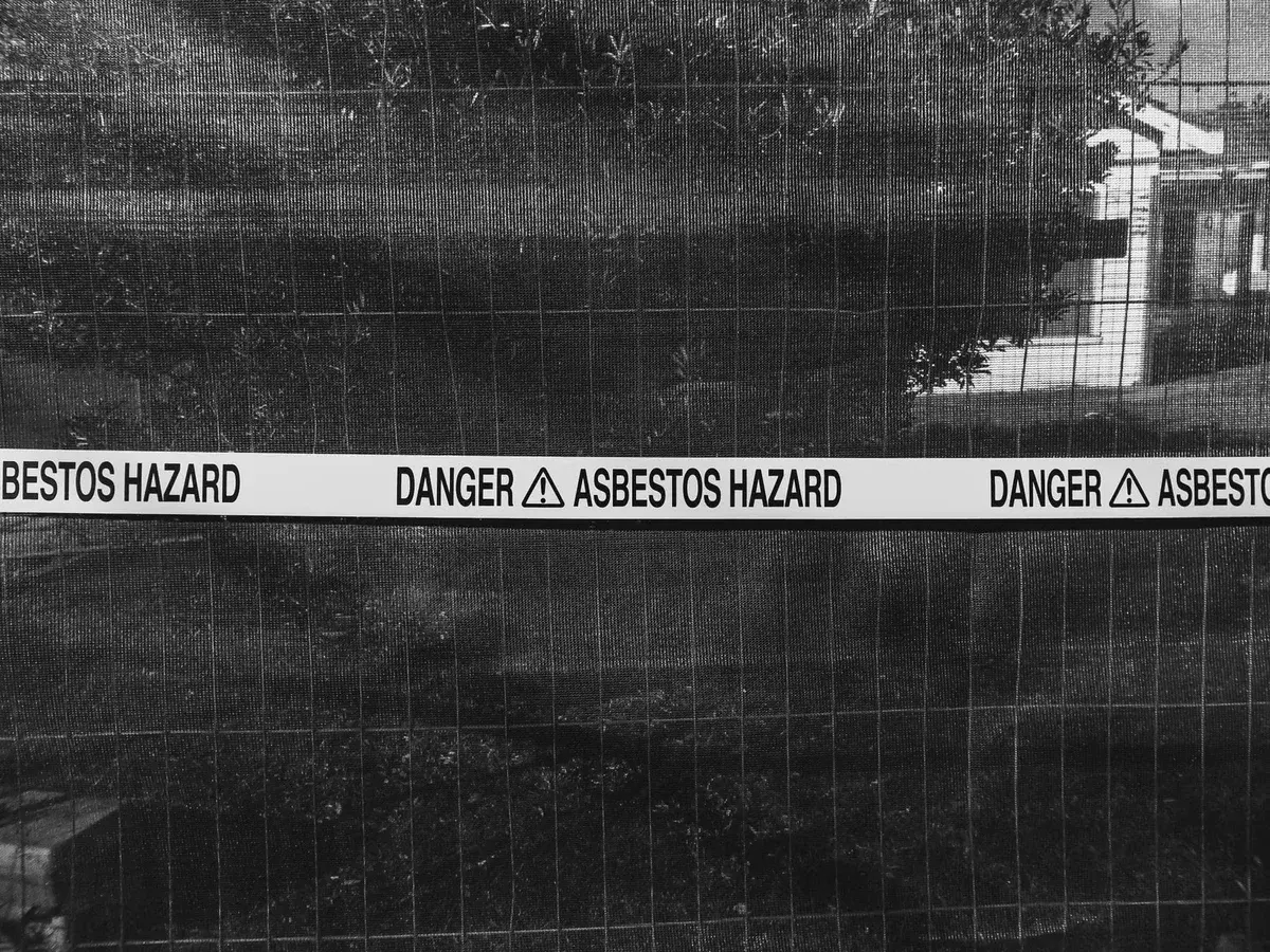 Black-and-white photo of a fence with asbestos hazard warning tape wrapped across it.
