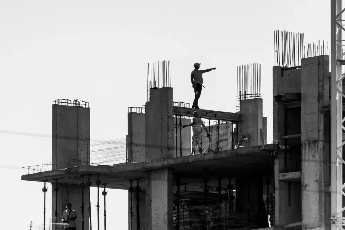 Silhouetted construction workers on an unfinished concrete building, one pointing outward.